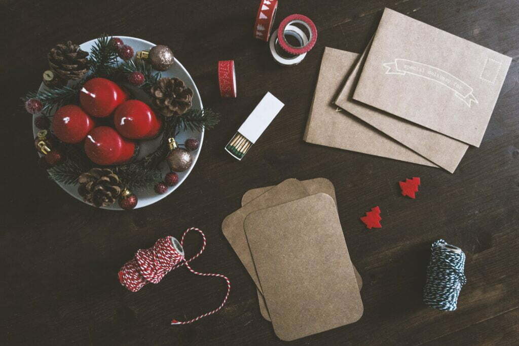 collection of christmas crafts on dark wooden table, including postcards, envelopes, christmas-themed string and tape, and a bowl of four red candles with pinecones and baubles placed around them, for christmas marketing planning article