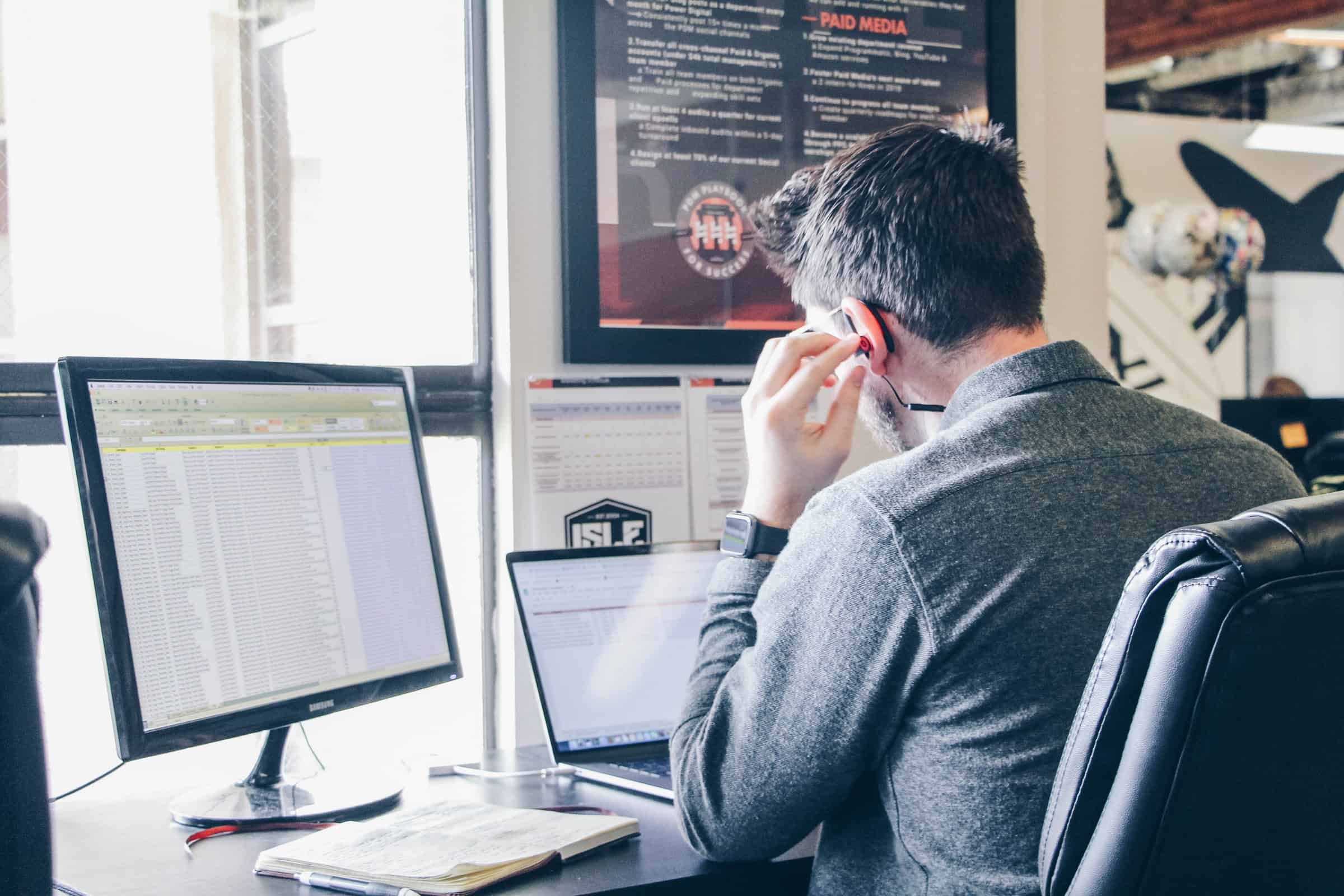 man sitting at desk with commputer and laptop listening to earphones for best salesman/salesperson article