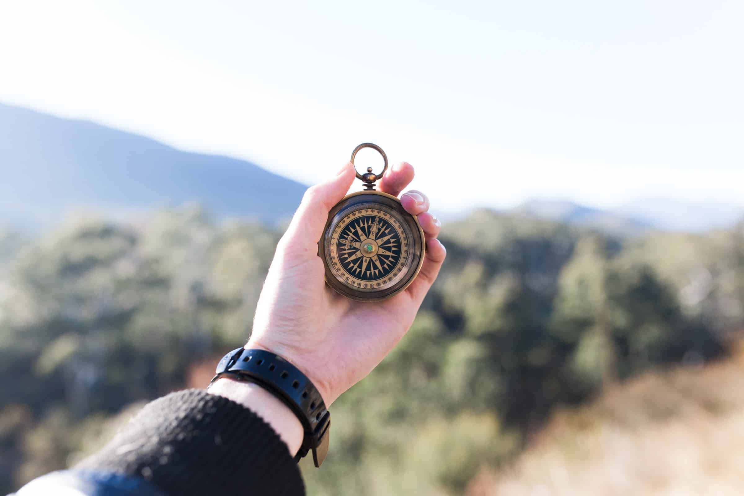 person holding compass in centre of photo with out of focus mountains and trees in the background for follow ups article