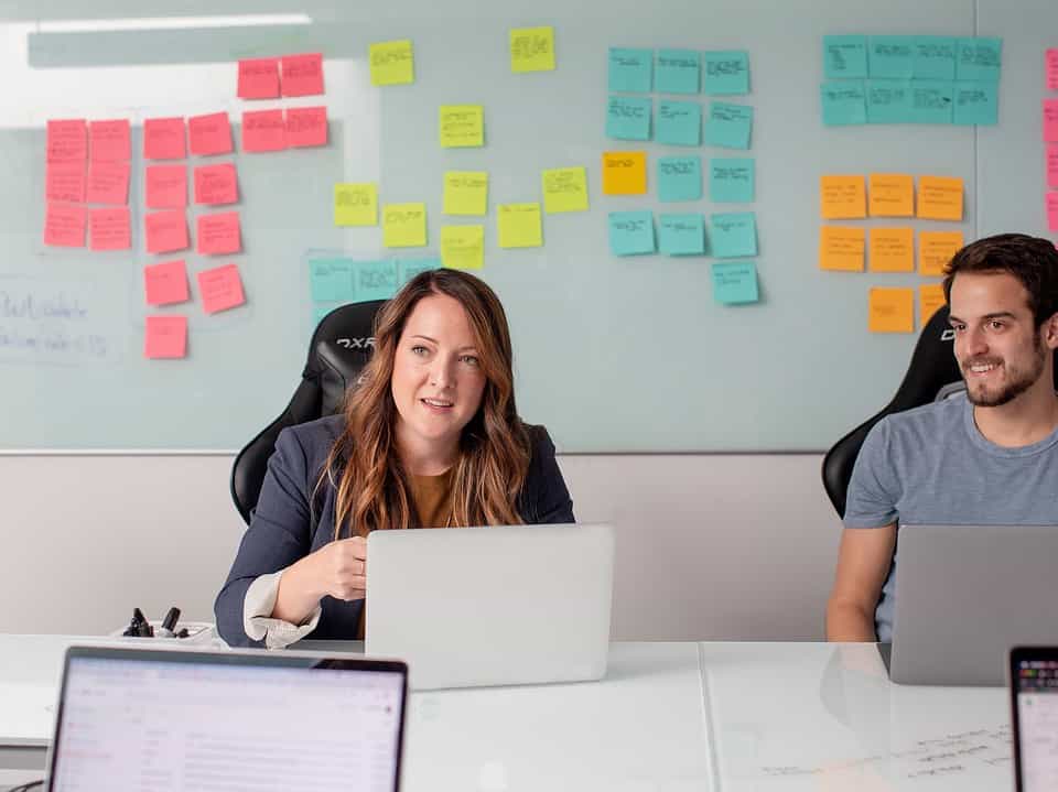 two people with laptops at business desk listening to person behind camera with board of sticky notes behind them for follow ups article