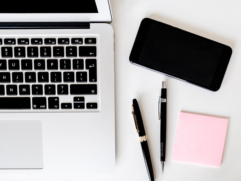 two pens, pink sticky notes and a smartphone near a macbook air pro on a white surface for adding a note hubspot vs popcorn comparison video