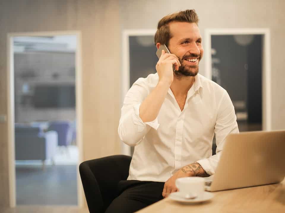 man on phone whilst sat at a wooden table with a macbook and coffee for retain customers article